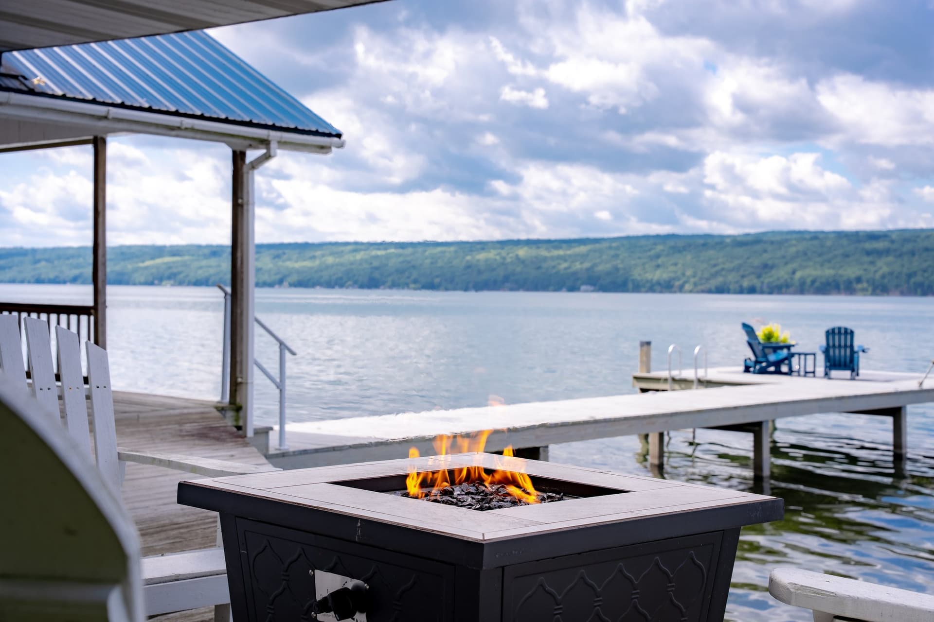 A lit gas fire table on the shore with flames burning, the dock running out past it into calm water, and the far shore low on the horizon under a wide cloudy sky.