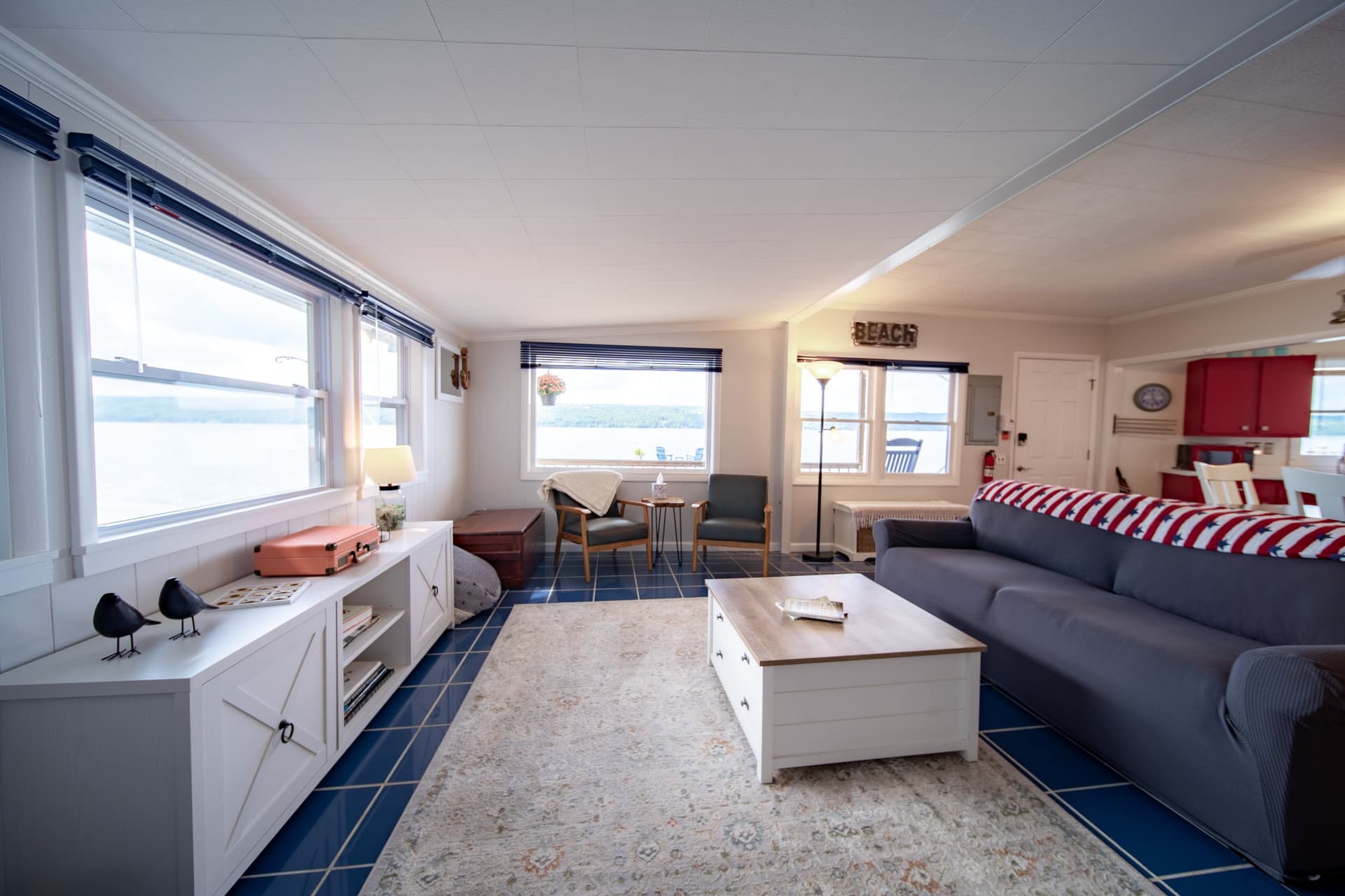 The living room with lake showing through every window along two walls, a grey sofa and white coffee table on a patterned rug over deep blue tile, and a pair of chairs in the far corner facing the water.