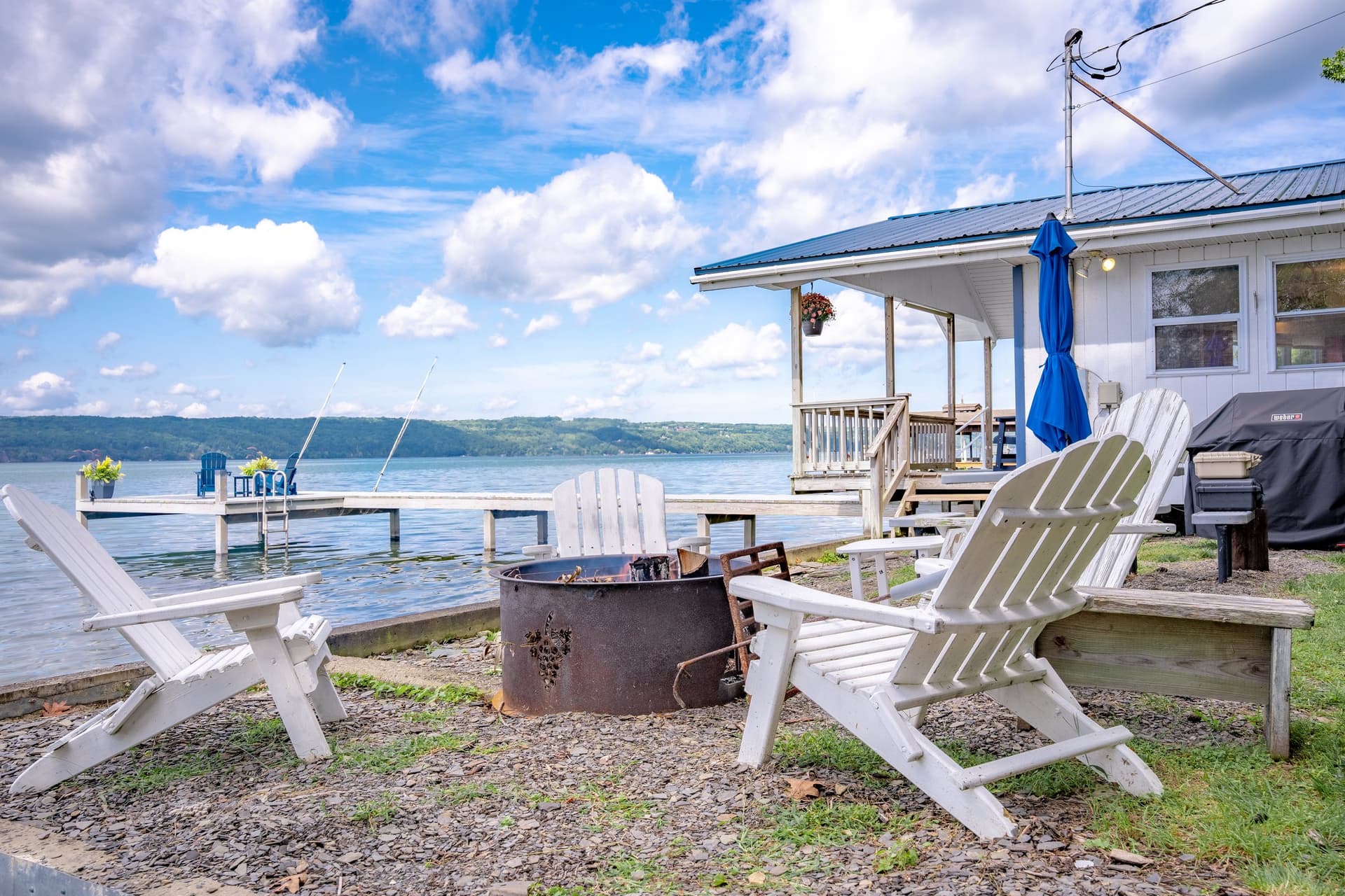 The shore terrace with white Adirondack chairs around a fire ring on the shingle, the cottage and its porch to the right, and the dock reaching into the lake behind.
